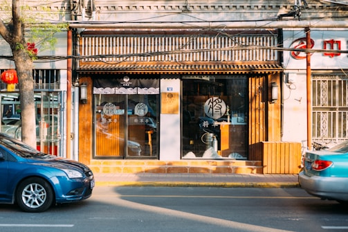 A storefront with a traditional Chinese design features a wooden facade and glass doors with decorative patterns. A blue car is parked on the street in front of the store. There is a tree with green leaves on the sidewalk and a red lantern hanging from it. The interior of the store is dimly visible, with some items on display.