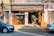 A storefront with a traditional Chinese design features a wooden facade and glass doors with decorative patterns. A blue car is parked on the street in front of the store. There is a tree with green leaves on the sidewalk and a red lantern hanging from it. The interior of the store is dimly visible, with some items on display.