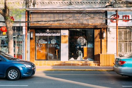 A storefront with a traditional Chinese design features a wooden facade and glass doors with decorative patterns. A blue car is parked on the street in front of the store. There is a tree with green leaves on the sidewalk and a red lantern hanging from it. The interior of the store is dimly visible, with some items on display.