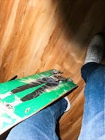 A light wood floor with a skateboard featuring a green design and a graphic. Someone wearing jeans and white sneakers is standing next to the skateboard, with one foot slightly above it.