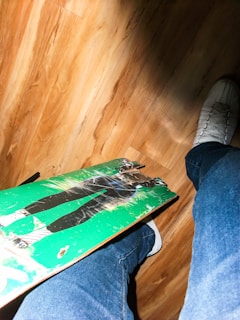 A light wood floor with a skateboard featuring a green design and a graphic. Someone wearing jeans and white sneakers is standing next to the skateboard, with one foot slightly above it.