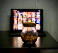 A glass jar filled with popcorn is placed on a dark wooden table. In the background, there's a blurred image of a tablet or screen displaying a grid of colorful thumbnails, suggesting a streaming service interface.