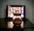 A glass jar filled with popcorn is placed on a dark wooden table. In the background, there's a blurred image of a tablet or screen displaying a grid of colorful thumbnails, suggesting a streaming service interface.