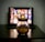 A glass jar filled with popcorn is placed on a dark wooden table. In the background, there's a blurred image of a tablet or screen displaying a grid of colorful thumbnails, suggesting a streaming service interface.