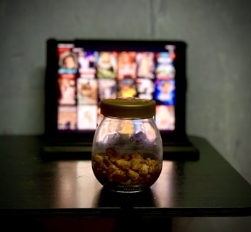 A glass jar filled with popcorn is placed on a dark wooden table. In the background, there's a blurred image of a tablet or screen displaying a grid of colorful thumbnails, suggesting a streaming service interface.