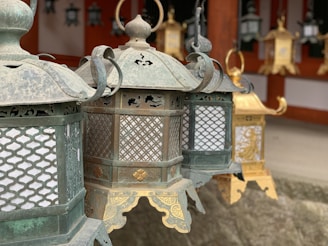 Several traditional lanterns with intricate metalwork hang in a line. The craftsmanship features delicate cut-out patterns and ornate details. The lanterns are primarily made of metal and exhibit a weathered patina, suggesting age or exposure to the elements. They are suspended against a backdrop that includes architectural elements.