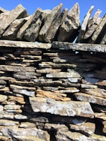 A close-up of a stone wall constructed with irregularly shaped, roughly cut stones stacked on top of each other. The stones vary in color from light gray to tan, and there is visible moss or lichen growing on some surfaces. The top row of the wall has stones arranged vertically, giving a jagged appearance against the clear blue sky.