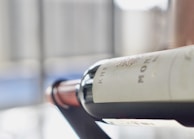 A wine bottle is resting on a metal rack at an angle, with a focus on the label that appears to have some text and an embossed emblem. The background is softly blurred, featuring vertical lines and hints of soft colors, creating a bokeh effect.