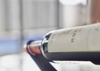 A wine bottle is resting on a metal rack at an angle, with a focus on the label that appears to have some text and an embossed emblem. The background is softly blurred, featuring vertical lines and hints of soft colors, creating a bokeh effect.