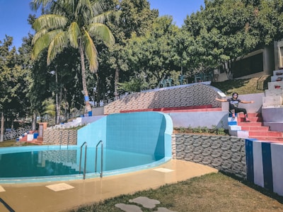 A circular swimming pool with turquoise water is surrounded by geometric tile patterns and a palm tree. There is a set of steps with red and blue tiles leading up to an area with trees and tables. A person is sitting on the steps with arms wide open, suggesting a relaxed and inviting atmosphere.