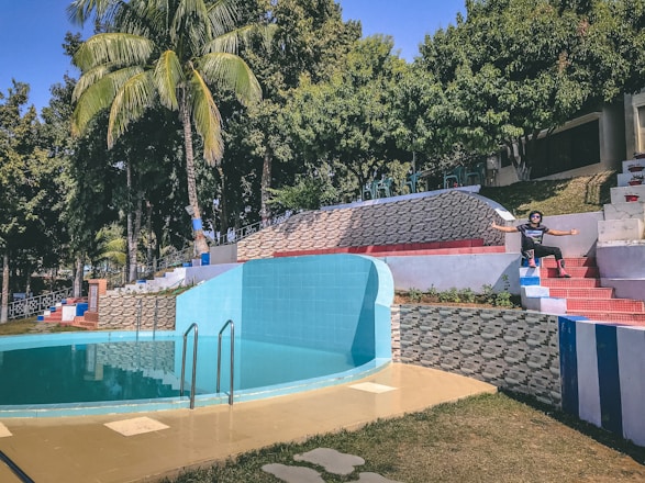 A circular swimming pool with turquoise water is surrounded by geometric tile patterns and a palm tree. There is a set of steps with red and blue tiles leading up to an area with trees and tables. A person is sitting on the steps with arms wide open, suggesting a relaxed and inviting atmosphere.