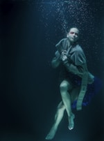 A person is submerged underwater with a serene expression. They are wrapped in a thick cloak or fabric with red makeup around their eyes. Bubbles gently rise towards the surface, adding to the tranquil and otherworldly atmosphere.