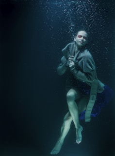 A person is submerged underwater with a serene expression. They are wrapped in a thick cloak or fabric with red makeup around their eyes. Bubbles gently rise towards the surface, adding to the tranquil and otherworldly atmosphere.