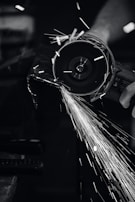 A close-up view of a person using a power grinder, generating bright sparks as it cuts through metal. The image is in black and white, highlighting the contrast between the dark environment and the bright, dynamic sparks flying through the air.