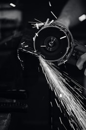 A close-up view of a person using a power grinder, generating bright sparks as it cuts through metal. The image is in black and white, highlighting the contrast between the dark environment and the bright, dynamic sparks flying through the air.