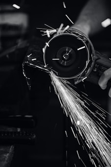 A close-up view of a person using a power grinder, generating bright sparks as it cuts through metal. The image is in black and white, highlighting the contrast between the dark environment and the bright, dynamic sparks flying through the air.