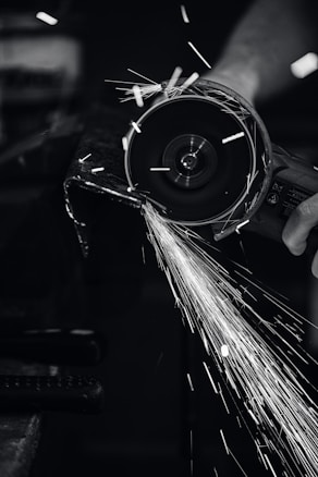 A close-up view of a person using a power grinder, generating bright sparks as it cuts through metal. The image is in black and white, highlighting the contrast between the dark environment and the bright, dynamic sparks flying through the air.
