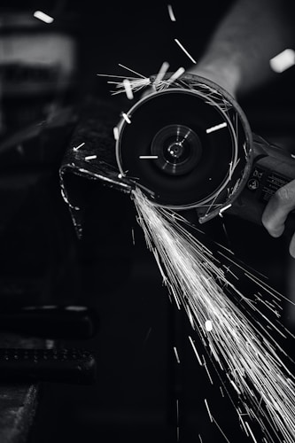 A close-up view of a person using a power grinder, generating bright sparks as it cuts through metal. The image is in black and white, highlighting the contrast between the dark environment and the bright, dynamic sparks flying through the air.