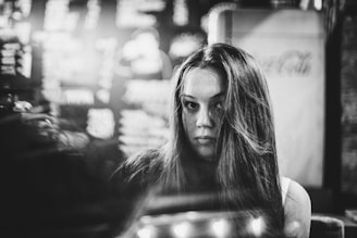 A black and white image featuring a woman with long hair. The background displays blurred text reminiscent of a menu or signage, suggesting a cafe or bar setting. There is a soft focus on the woman's face, giving the image a reflective and pensive quality.