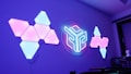 A futuristic wall features geometric-shaped light panels with a mix of blue and pink hues, arranged in triangular formations on both sides. In the center, a circular neon light with a stylized design glows brightly. The room has a modern aesthetic with a purple tint, enhancing the vibrant lighting effect.