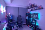 A modern gaming setup featuring a sleek gaming chair, dual monitors on a desk, and LED lighting. A gaming console and various decorations are displayed on shelves, creating an organized and vibrant gaming environment.