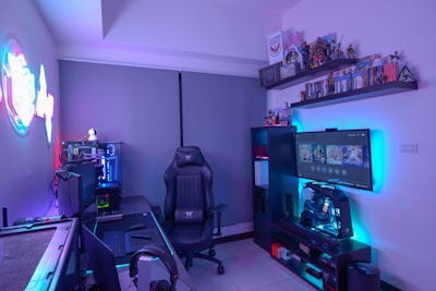 A modern gaming setup featuring a sleek gaming chair, dual monitors on a desk, and LED lighting. A gaming console and various decorations are displayed on shelves, creating an organized and vibrant gaming environment.