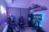 A modern gaming setup featuring a sleek gaming chair, dual monitors on a desk, and LED lighting. A gaming console and various decorations are displayed on shelves, creating an organized and vibrant gaming environment.