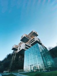 A modern architectural structure featuring a series of stacked, rectangular modules with a large glass facade in the foreground. The building is set against a clear blue sky, with trees visible at the base.