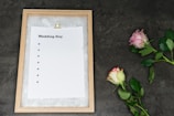 A wooden-framed board holds an empty checklist with the title 'Wedding Day,' featuring bullet points but no text. Two roses, one pink and one white with pink edges, lie on a dark gray textured surface beside the board.