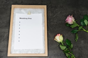 A wooden-framed board holds an empty checklist with the title 'Wedding Day,' featuring bullet points but no text. Two roses, one pink and one white with pink edges, lie on a dark gray textured surface beside the board.