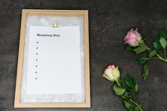 A wooden-framed board holds an empty checklist with the title 'Wedding Day,' featuring bullet points but no text. Two roses, one pink and one white with pink edges, lie on a dark gray textured surface beside the board.