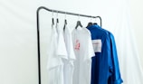 Several white and blue shirts are hung on a black metal clothing rack against a light backdrop. The white shirts feature red graphic designs, and a contrasting blue shirt is placed next to them.