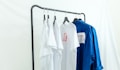 Several white and blue shirts are hung on a black metal clothing rack against a light backdrop. The white shirts feature red graphic designs, and a contrasting blue shirt is placed next to them.