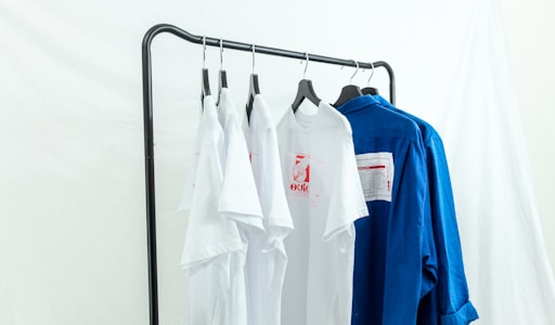 Several white and blue shirts are hung on a black metal clothing rack against a light backdrop. The white shirts feature red graphic designs, and a contrasting blue shirt is placed next to them.