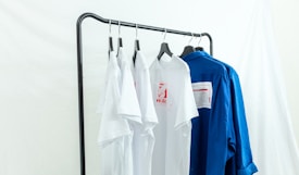 Several white and blue shirts are hung on a black metal clothing rack against a light backdrop. The white shirts feature red graphic designs, and a contrasting blue shirt is placed next to them.