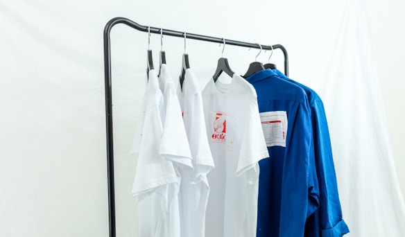 Several white and blue shirts are hung on a black metal clothing rack against a light backdrop. The white shirts feature red graphic designs, and a contrasting blue shirt is placed next to them.