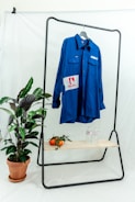 A bright blue button-up shirt is hanging on a black metal rack against a plain white backdrop. The shirt has patches with logos and text. Below the shirt, there is a wooden shelf holding two bottles of water and several ripe tomatoes with leaves. To the side, a potted plant with green leaves sits on the floor.