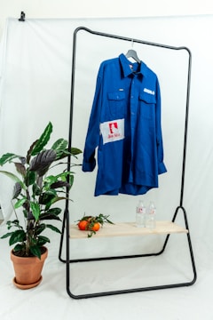 A bright blue button-up shirt is hanging on a black metal rack against a plain white backdrop. The shirt has patches with logos and text. Below the shirt, there is a wooden shelf holding two bottles of water and several ripe tomatoes with leaves. To the side, a potted plant with green leaves sits on the floor.