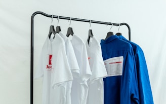 A black clothing rack holds four garments on hangers. Three of the items are white t-shirts with red graphics, and the last one is a blue shirt with a label on the back. The background is a plain white, creating a clean and minimalist look.