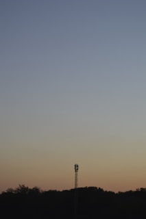 A lone communication tower stands silhouetted against a gradient sky transitioning from a deep blue at the top to soft shades of orange near the horizon. The horizon itself features a line of dark, silhouetted trees.