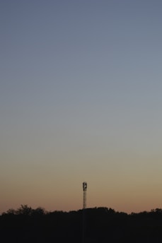 A lone communication tower stands silhouetted against a gradient sky transitioning from a deep blue at the top to soft shades of orange near the horizon. The horizon itself features a line of dark, silhouetted trees.