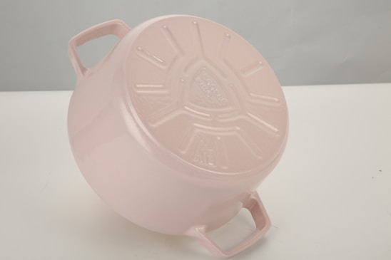 A light pink cast iron pot is placed upside down on a light-colored surface. The base of the pot features embossed text and a set of design patterns, with two handles visible protruding from the sides.