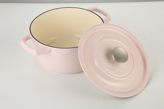 A pale pink cookware pot with two handles and a matching lid resting on its side. The interior of the pot is cream-colored, and the lid has a metal knob. The pot is placed on a smooth, white surface.