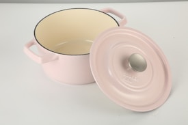 A pale pink cookware pot with two handles and a matching lid resting on its side. The interior of the pot is cream-colored, and the lid has a metal knob. The pot is placed on a smooth, white surface.