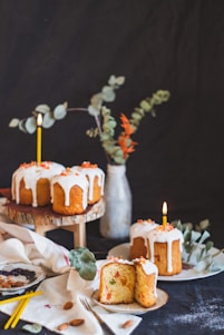 Several round cakes with white icing and orange sprinkles are displayed on a table. Some cakes are on a wooden stand, while others are on plates. Each cake has a lit candle. There are decorative elements including a vase with green and orange leaves, a white cloth, almonds, and a plate with remnants of food. The background is dark.