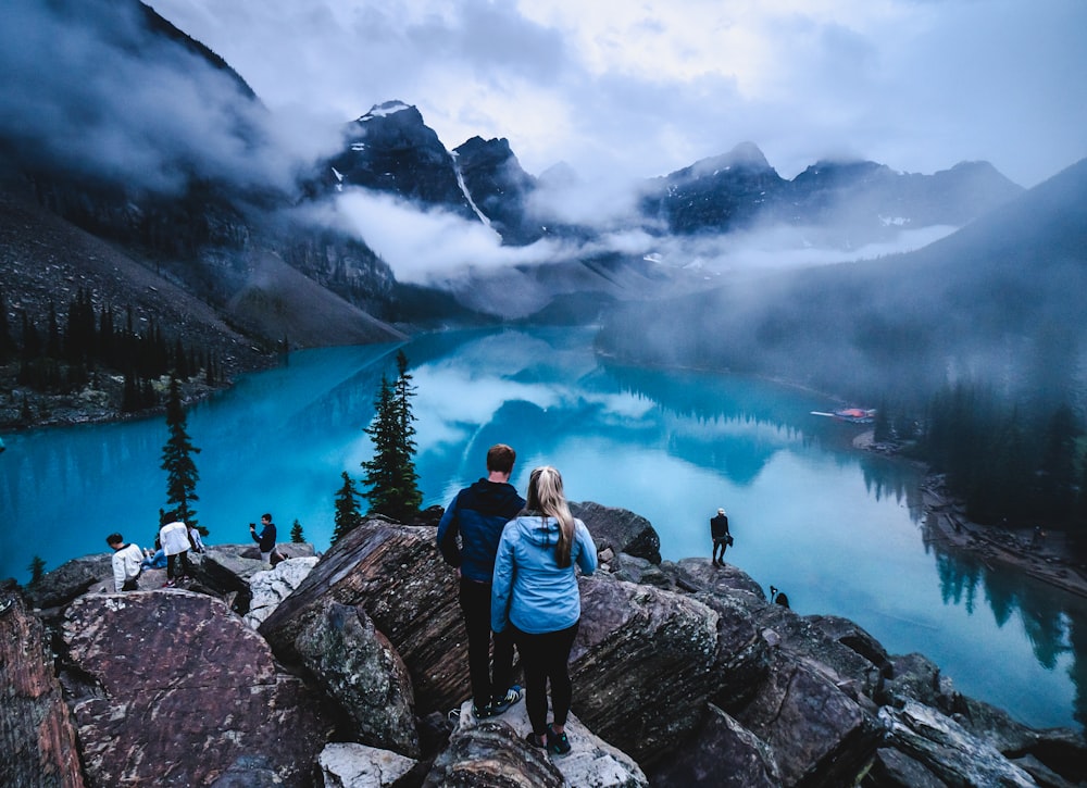 man and woman sitting on rock near lake under white clouds during daytime