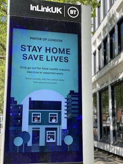 A public information screen installed by InLinkUK, featuring a COVID-19 safety message from the Mayor of London. The sign advises staying home to save lives, allowing only necessary outings for food, health reasons, exercise, or essential work. The illustration includes a house with individuals visible in the windows, surrounded by city buildings.