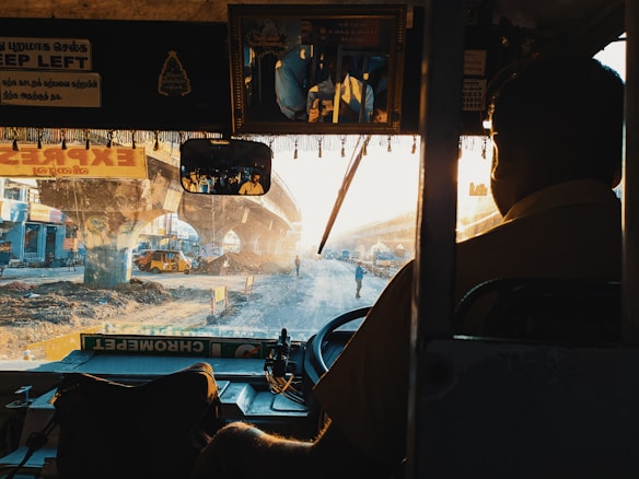 A view from inside a bus, with a driver wearing a uniform visible in the foreground. The outside scene shows a busy urban setting with a partially constructed overpass, dirt and construction equipment. Sunlight streams through the scene, creating a warm, hazy atmosphere. There are several signs inside the bus, one in English reading 'Keep Left'.