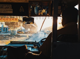 A view from inside a bus, with a driver wearing a uniform visible in the foreground. The outside scene shows a busy urban setting with a partially constructed overpass, dirt and construction equipment. Sunlight streams through the scene, creating a warm, hazy atmosphere. There are several signs inside the bus, one in English reading 'Keep Left'.
