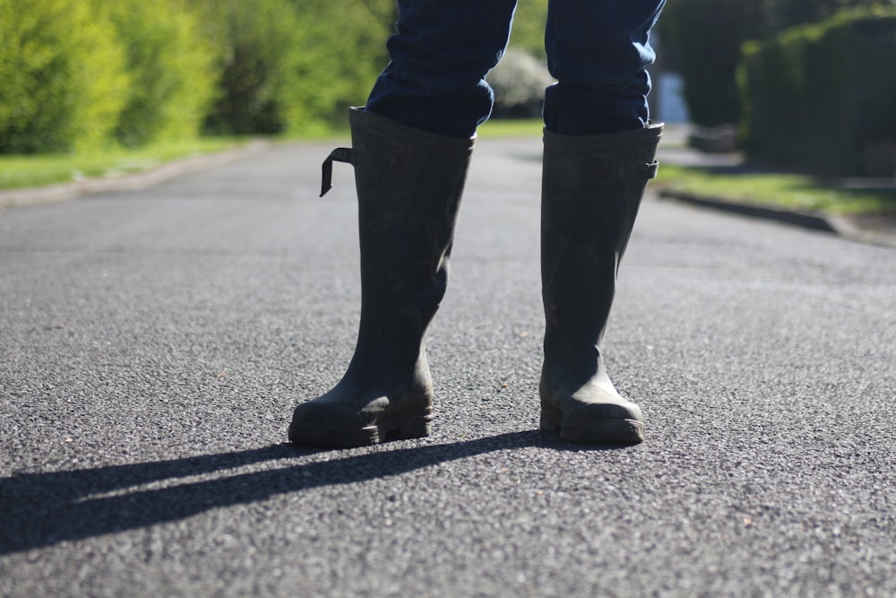 person in black rain boots standing on gray asphalt road during daytime